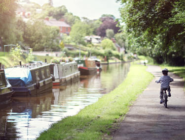 Narrowboats moored up on the side of a canal with young boy cycling on walk way